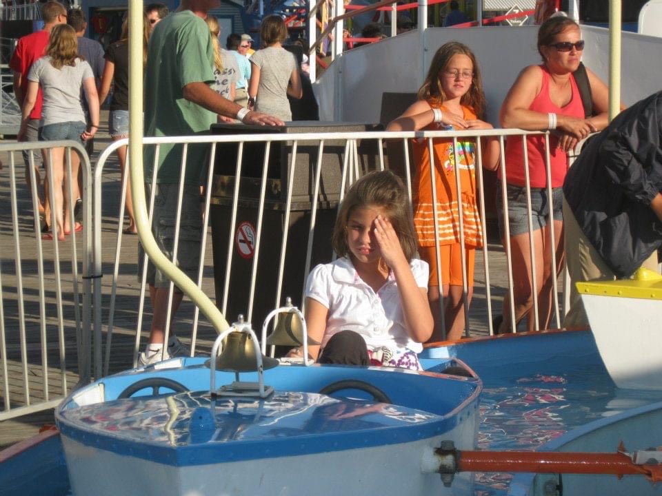 Young Jessi Walker sitting on an amusement park ride that is a boat ride seeming miserable from the sun light